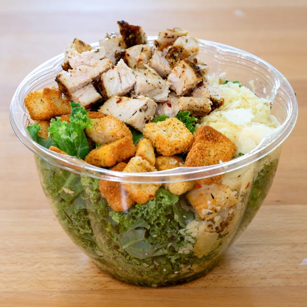 Fresh Green's Caesar salad with grilled chicken, croutons, and parmesan cheese in a clear plastic bowl.