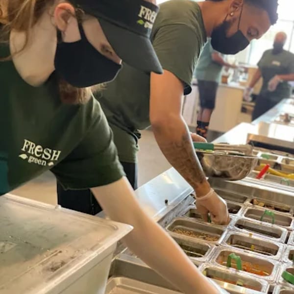 Fresh Green employees in masks and green shirts prepare salads at a food bar.