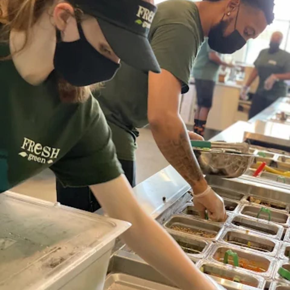 Fresh Green employees in masks and green shirts prepare salads at a food bar.