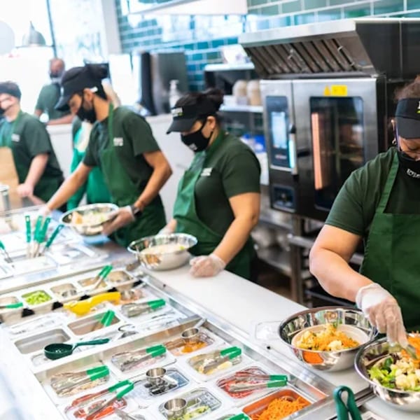 Fresh Green employees in green aprons and masks prepare salads behind a counter with various ingredients.