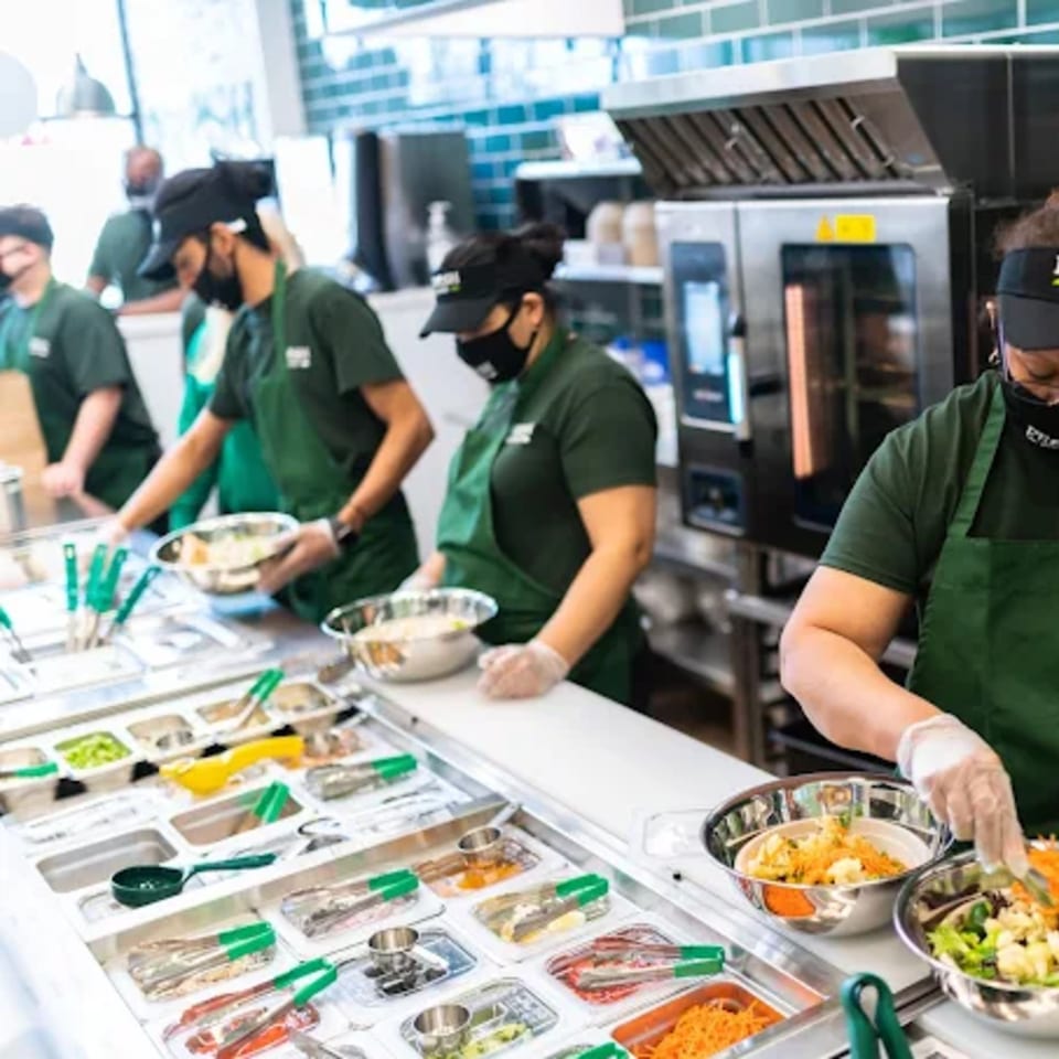 Fresh Green employees in green aprons and masks prepare salads behind a counter with various ingredients.