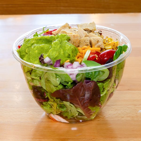 Fresh Green salad with guacamole, chicken, corn, tomatoes, and red onion in a clear plastic bowl.